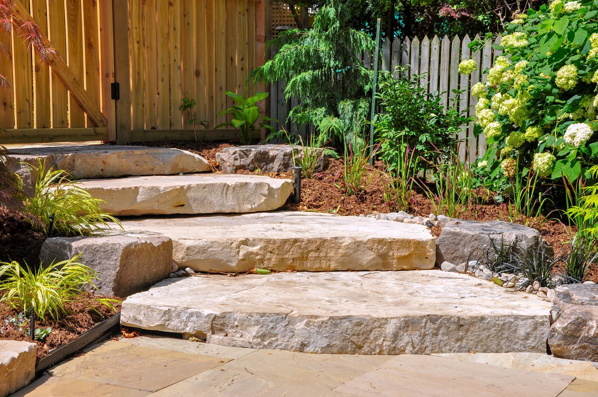 Large stone steps set in a garden with a wooden fence and flowering shrubs in the background.