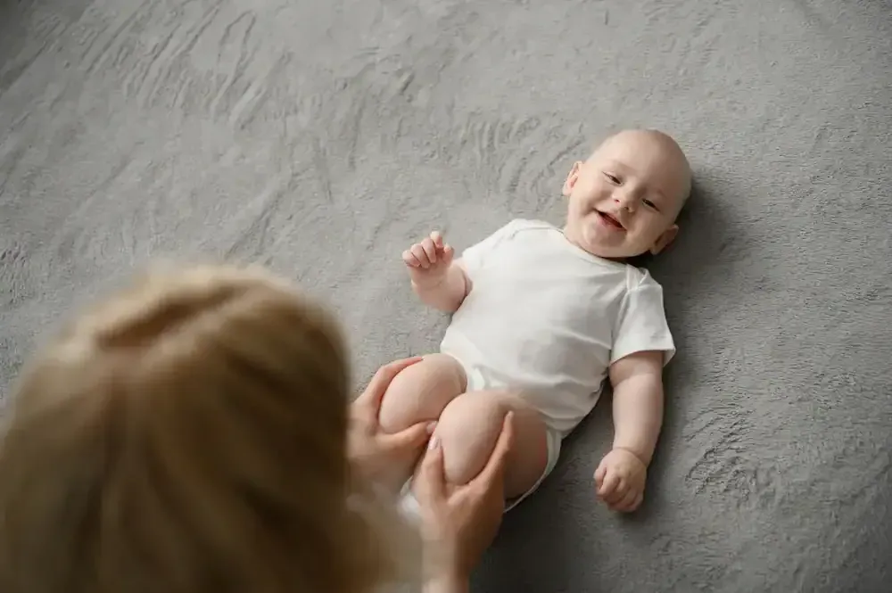 Smiling baby in white onesie lying on gray blanket, being touched by an adult