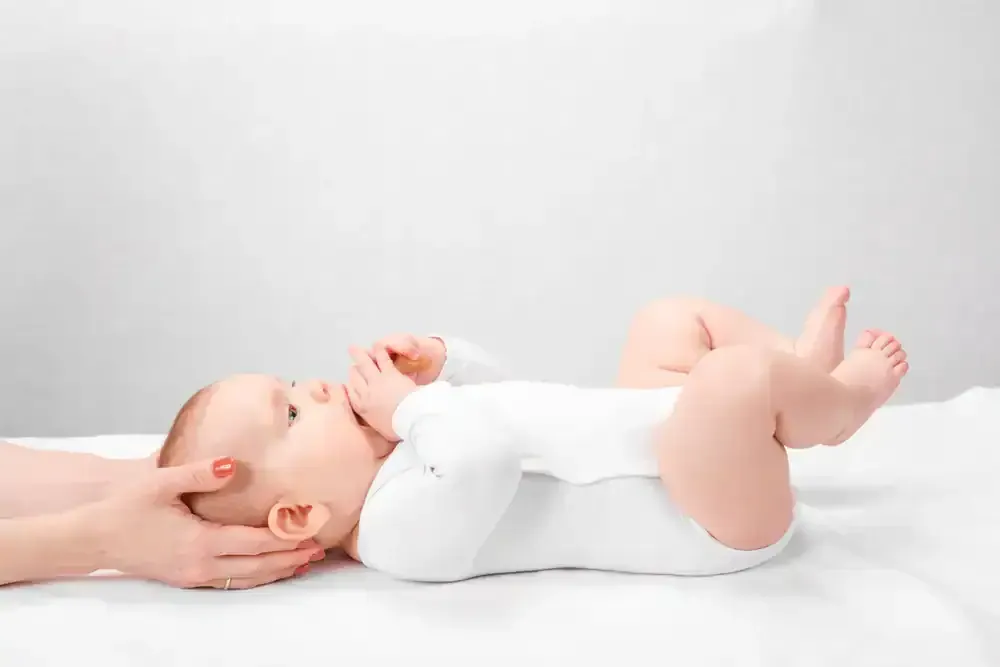 Baby lying on a white blanket, wearing a white onesie and holding a hand near the face.