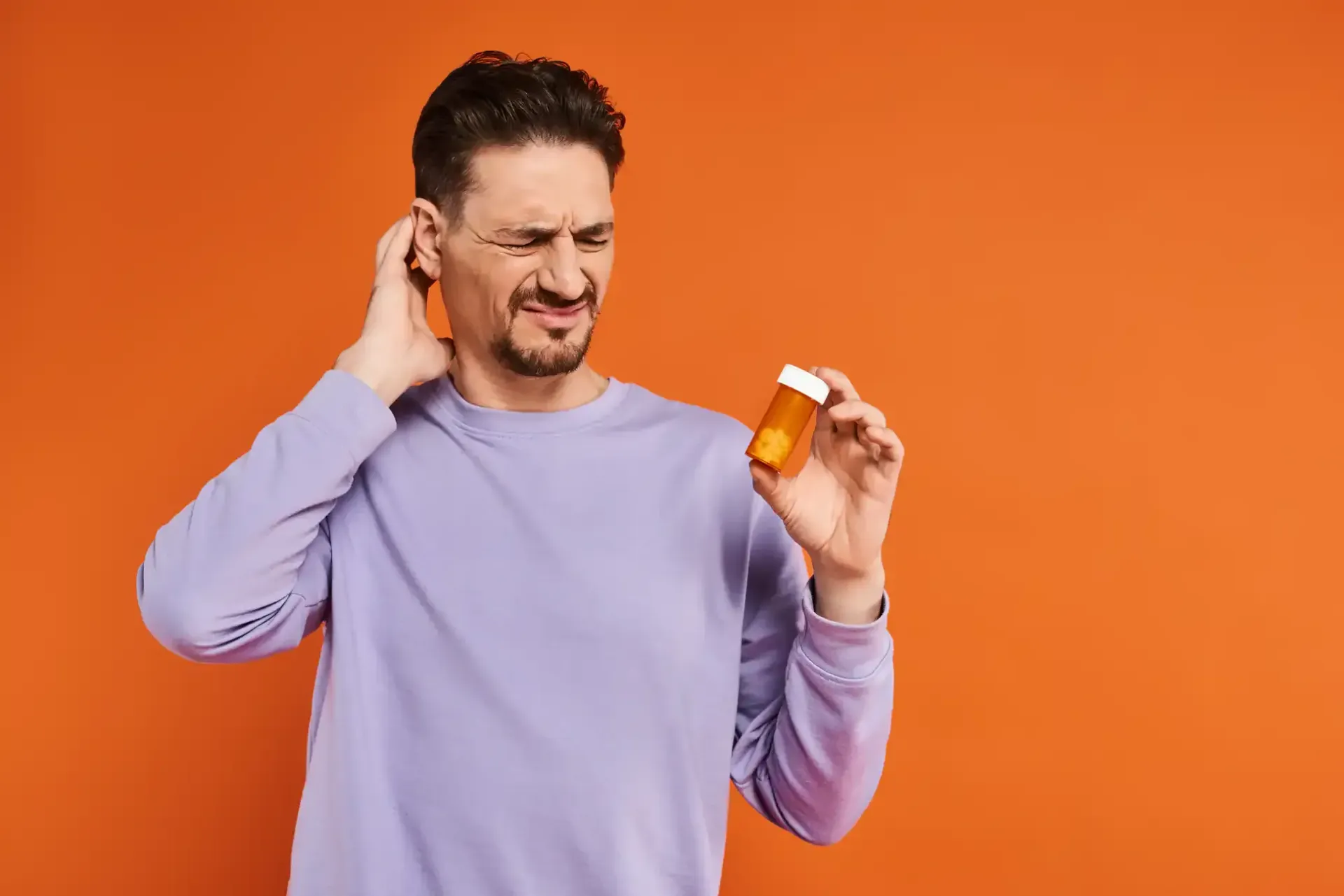 Man grimacing and holding an orange pill bottle against an orange background