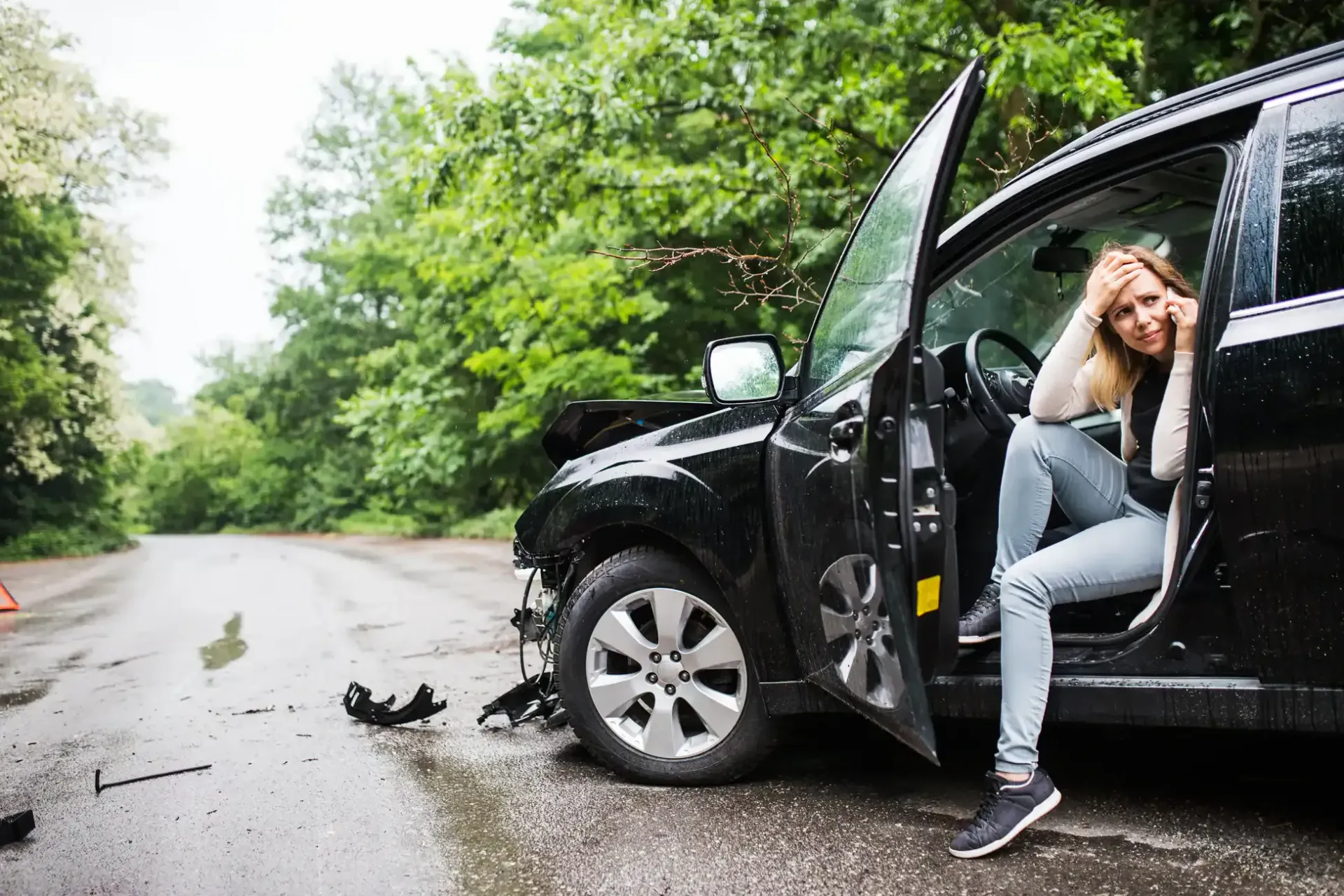 Woman stands by open car door after roadside crash on wet road with debris and damaged black vehicle