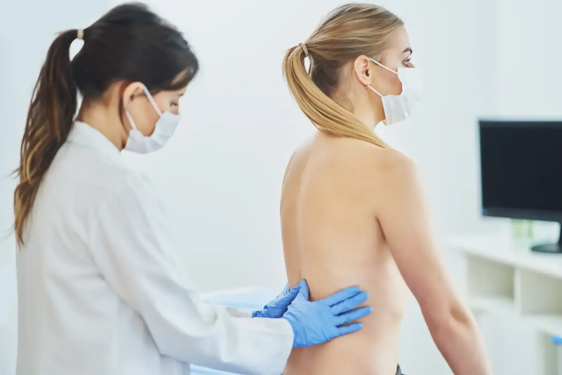 Healthcare worker examining a patient’s back in a clinic, both wearing masks and gloves