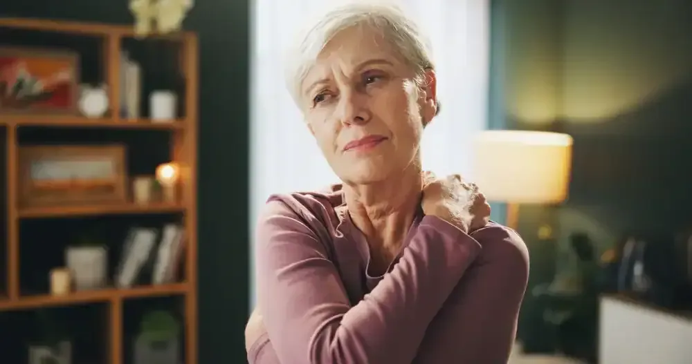 Woman in a cozy room, gently holding her shoulder with a thoughtful expression beside a lit lamp.