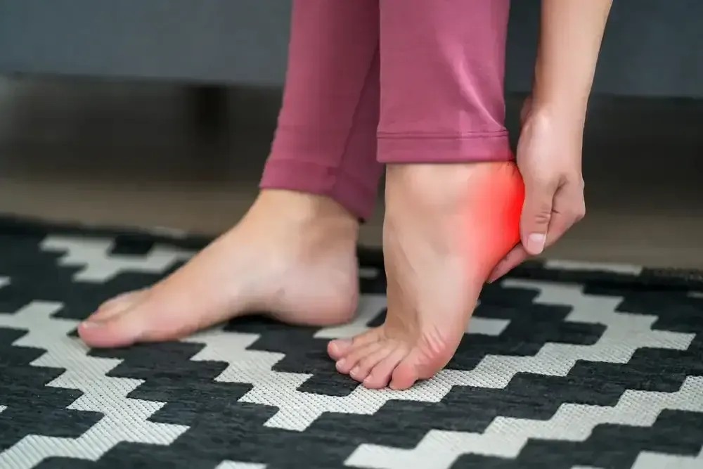 Person rubbing an ankle with a red pain highlight while standing on a patterned rug