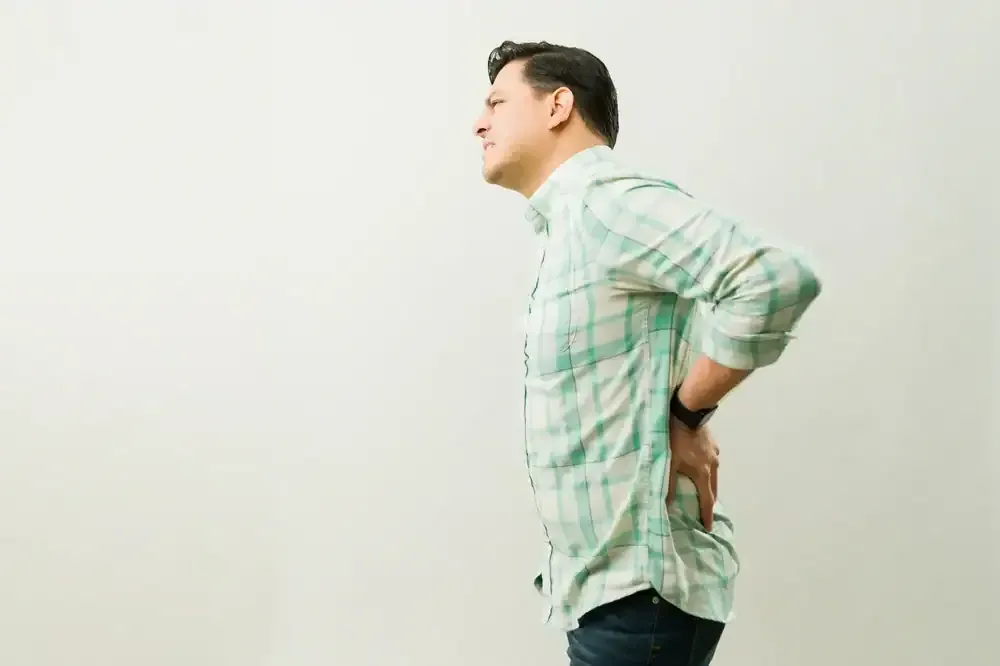 Side view of a man in a light green plaid shirt holding his lower back against a plain white background