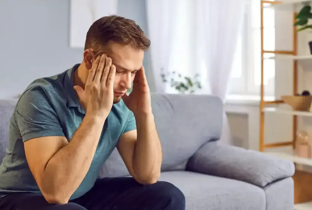 Man sitting on a gray sofa, holding his head with both hands in a bright living room