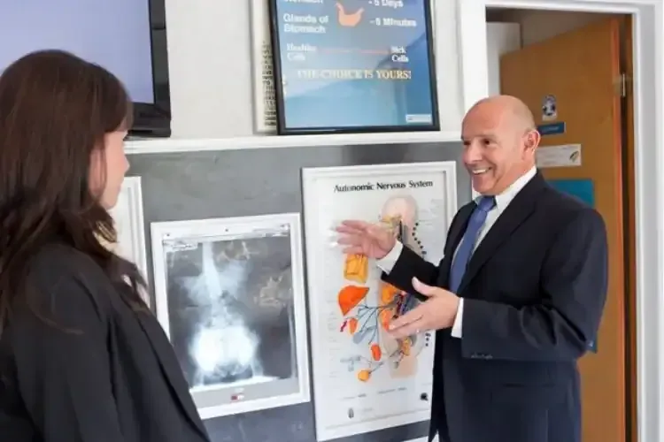 Man in a suit pointing at a medical poster while speaking with a woman in a clinic hallway