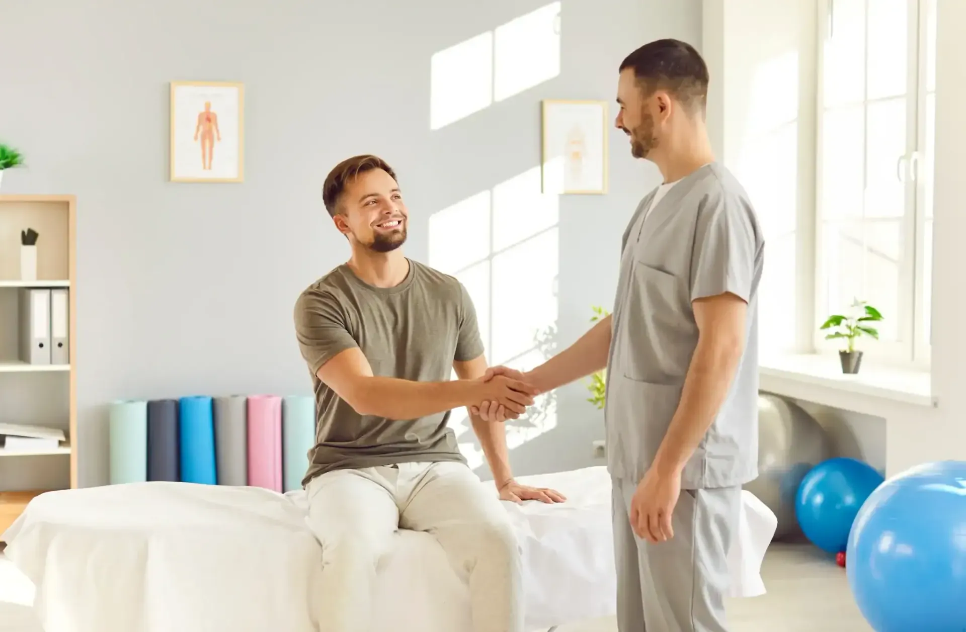 Two people shaking hands in a bright therapy room with exercise balls and a bed nearby