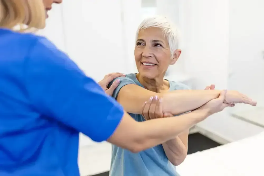 Woman in blue shirt stretching arm with a trainer in a bright gym setting