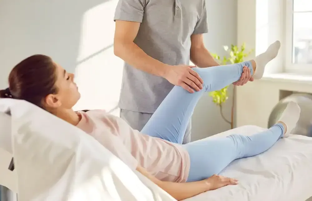 Caregiver helping a patient stretch her leg on a bed in a bright room