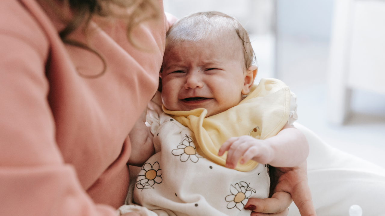 Crying baby in a yellow bib being held by an adult in a bright room