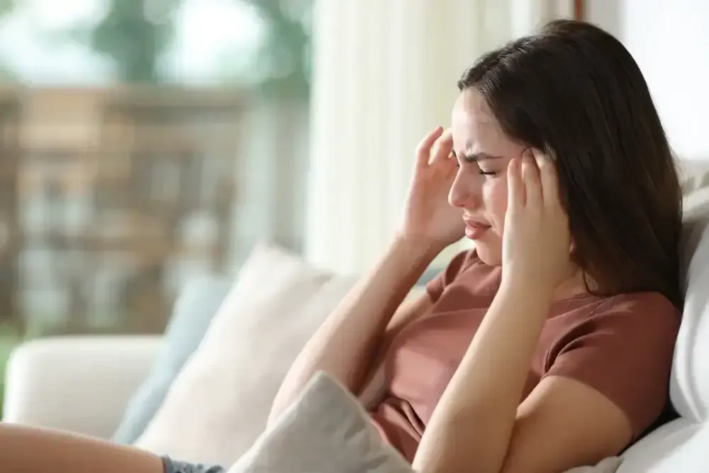 Woman sitting on a couch with eyes closed and hands at her temples, looking stressed.