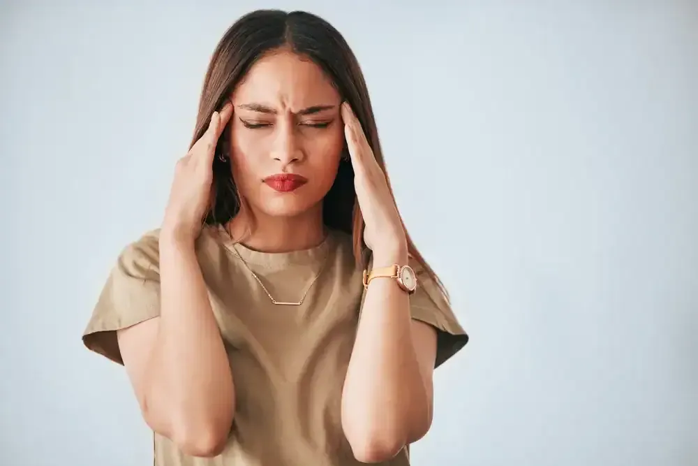 Woman in a beige shirt pressing her temples, eyes closed, showing a headache or stress.