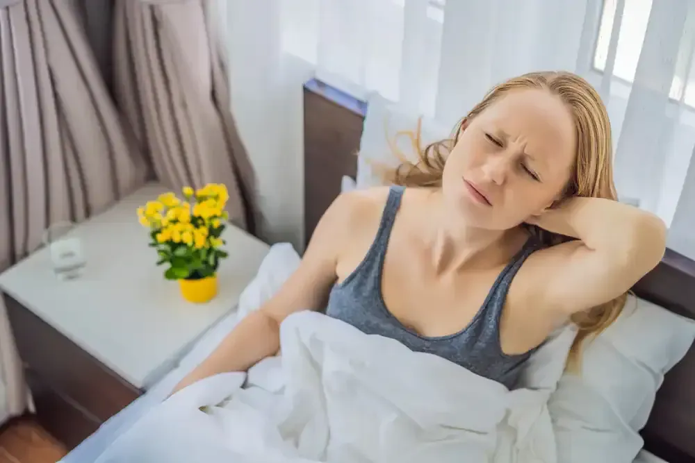 Woman resting in bed with a hand on her forehead, appearing unwell, beside a small table with yellow flowers.