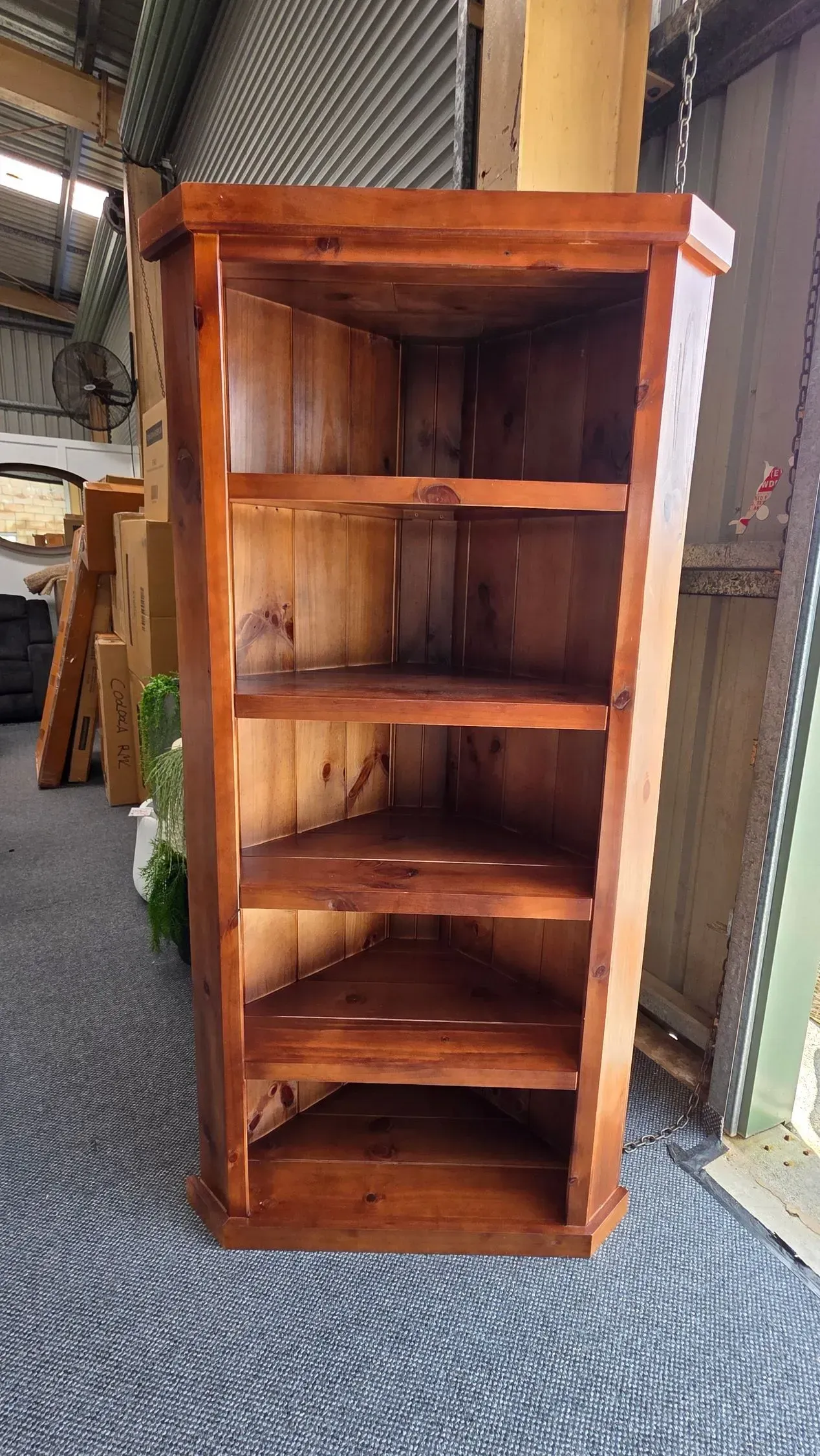 Wooden corner bookshelf with four shelves, shown indoors on a patterned rug.