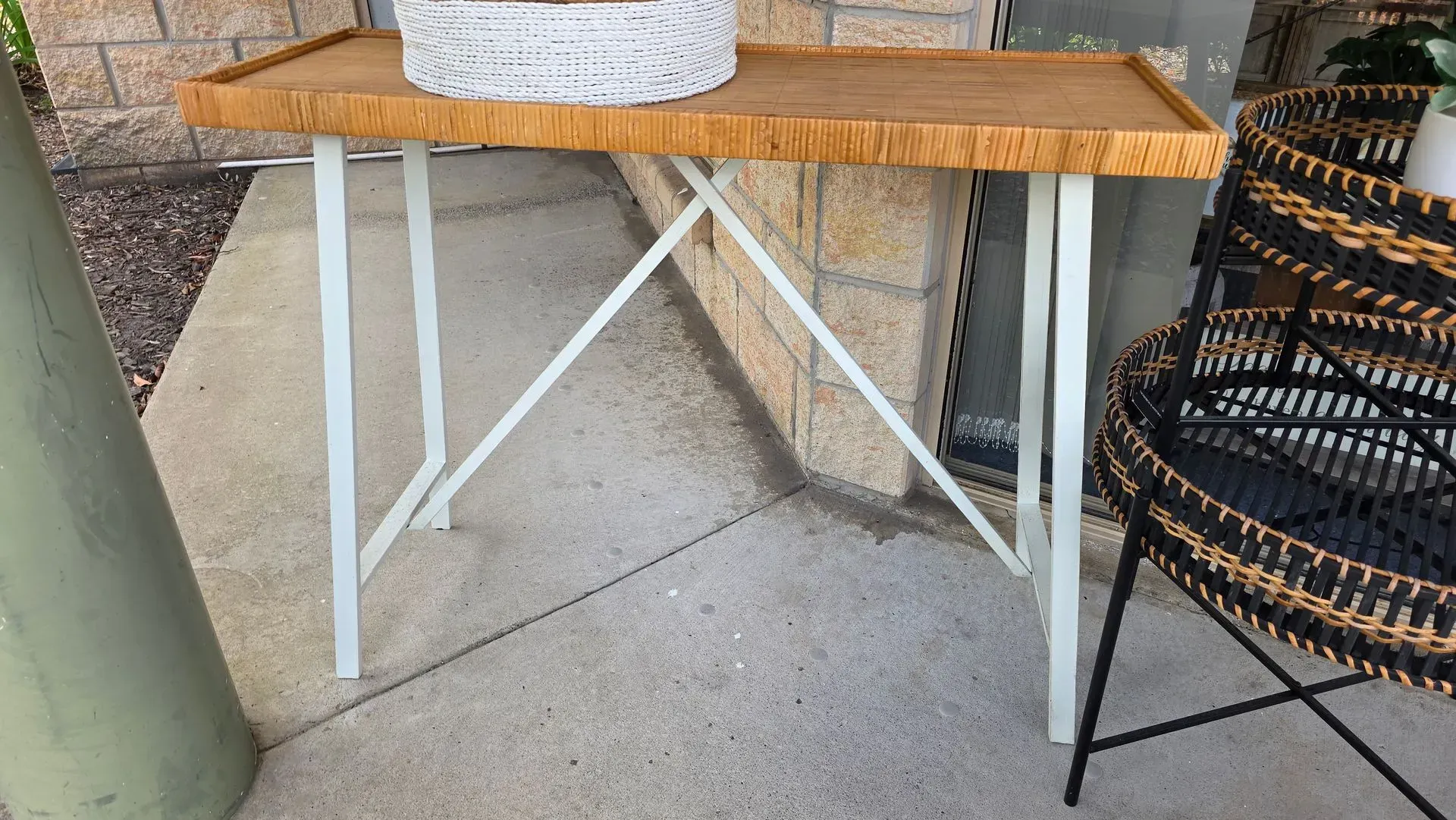 Rustic wooden table with white metal legs on a patio, beside a black woven chair and a green post