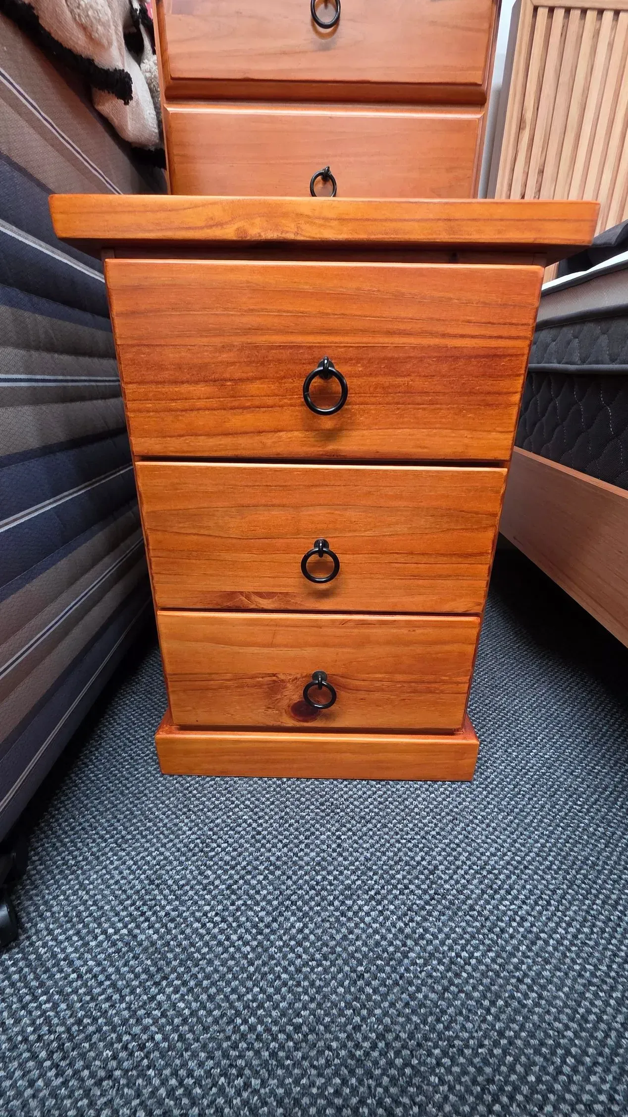 Small wooden three-drawer bedside table with brass pulls on a carpeted floor between two beds.