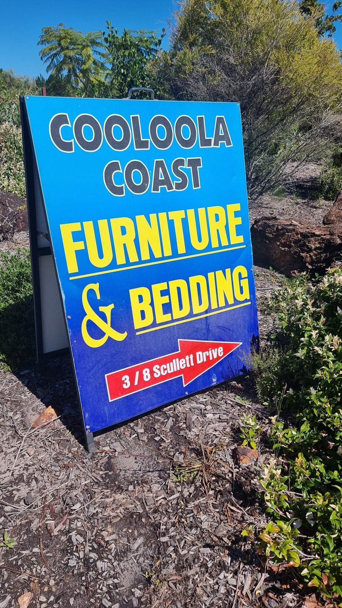 Blue sign for Cooloola Coast Furniture & Bedding in a garden bed, with trees and shrubs behind it.
