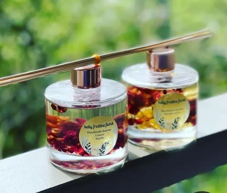 Two perfume bottles on a ledge, with gold caps and dried flowers inside, against a blurred green background