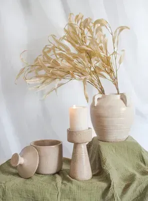 Neutral-toned ceramic vases and candle on a draped green cloth with dried beige stems against a light backdrop