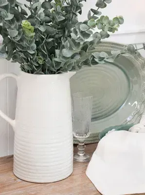 White vase with eucalyptus branches beside stacked green glass plates on a wooden table