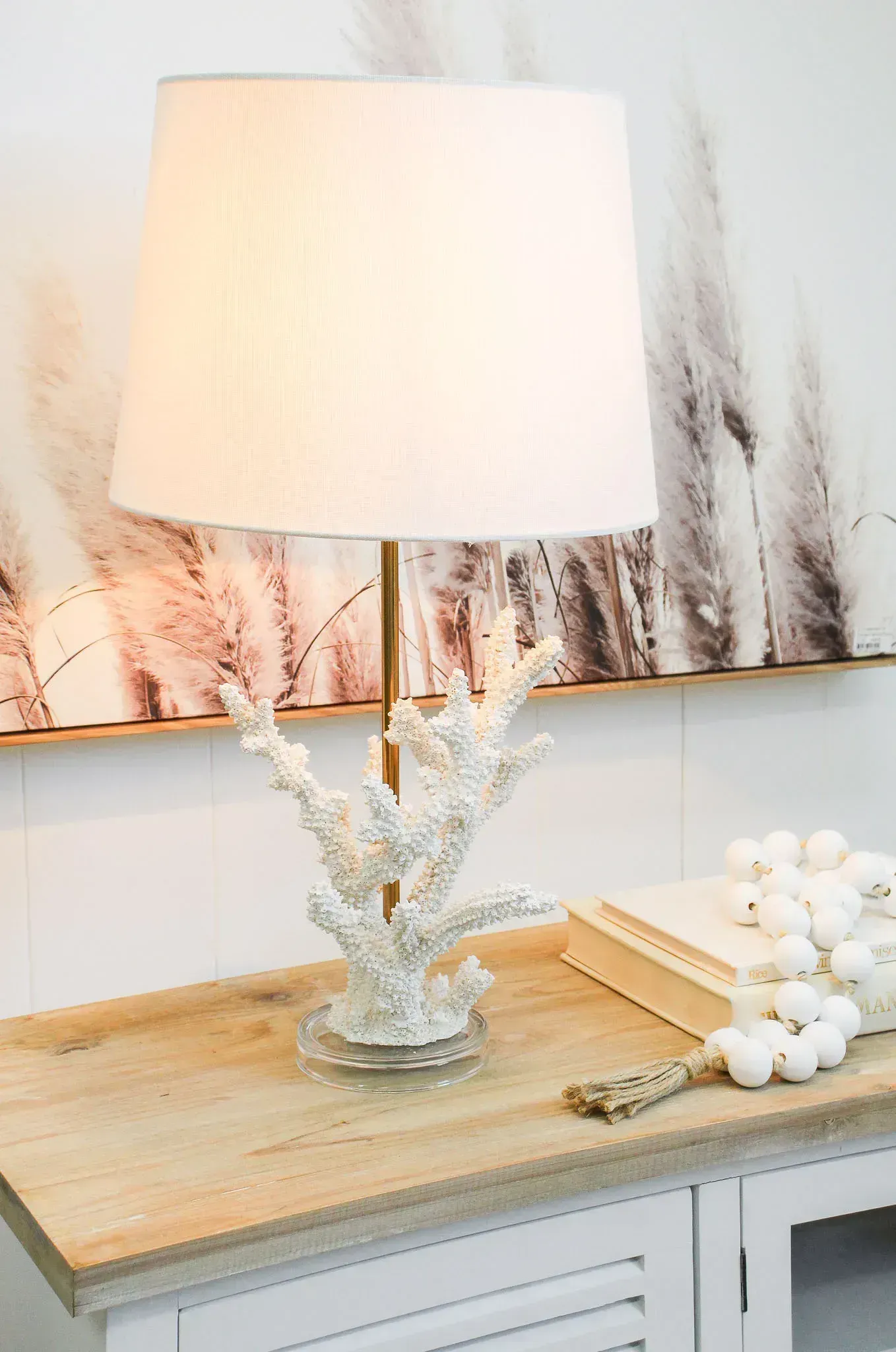 White table lamp on a wooden tray atop a distressed white console with coral decor and books