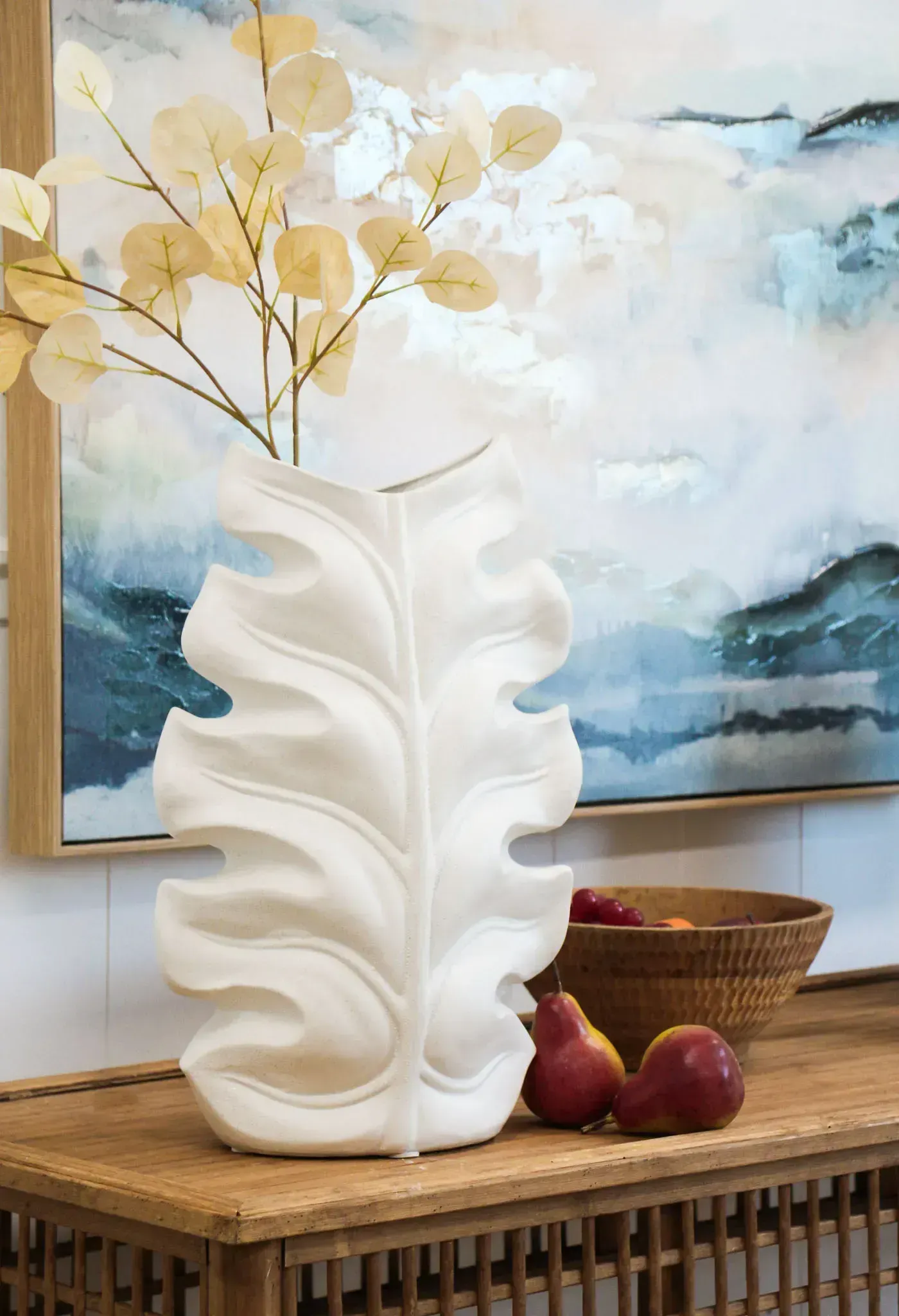 White leaf-shaped vase with dried flowers on a wooden table, with fruit and a basket in the background