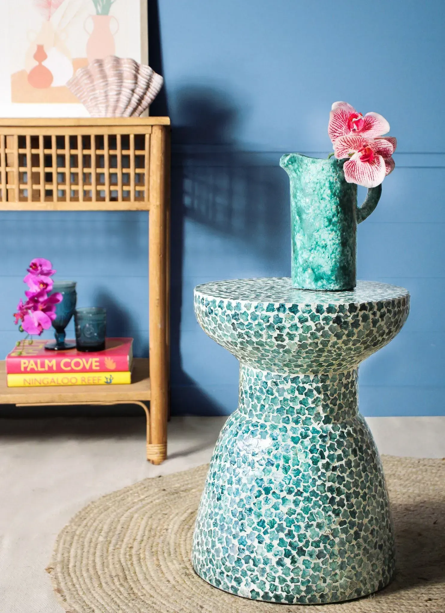 Turquoise mosaic stool with flowers beside a wooden shelf on a blue wall and woven rug.