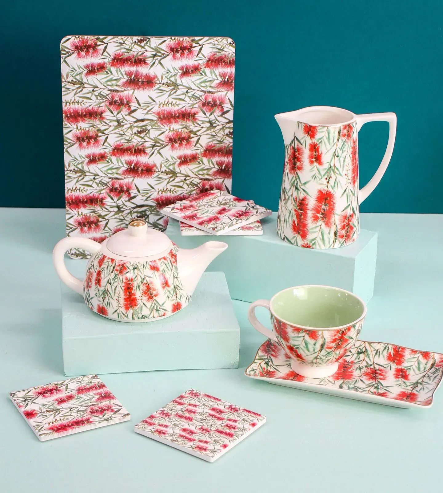 Floral red-and-green tea set with teapot, pitcher, cups, and coasters on pale blue blocks