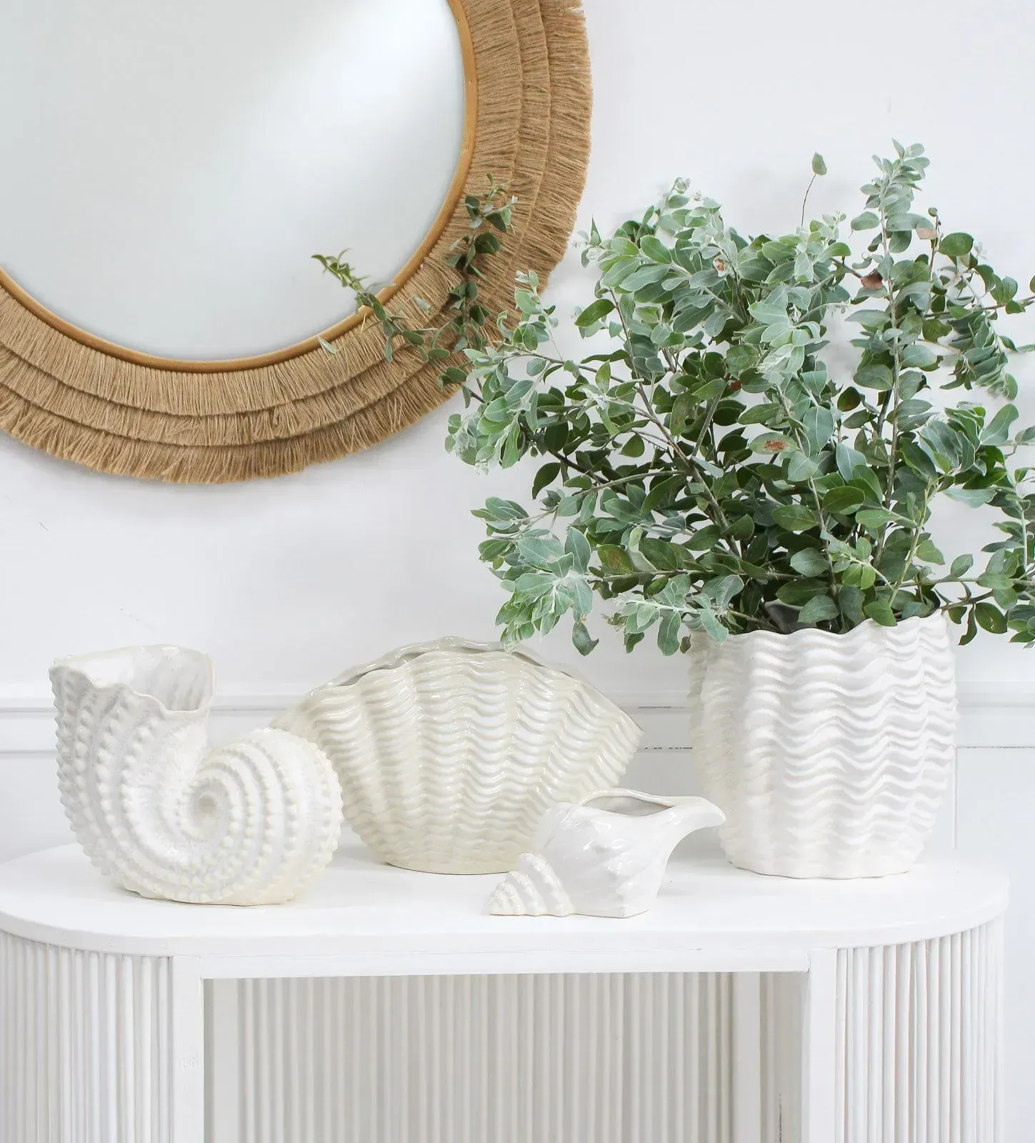 White side table with woven decor, sculptural vases, and a leafy potted plant against a white wall.