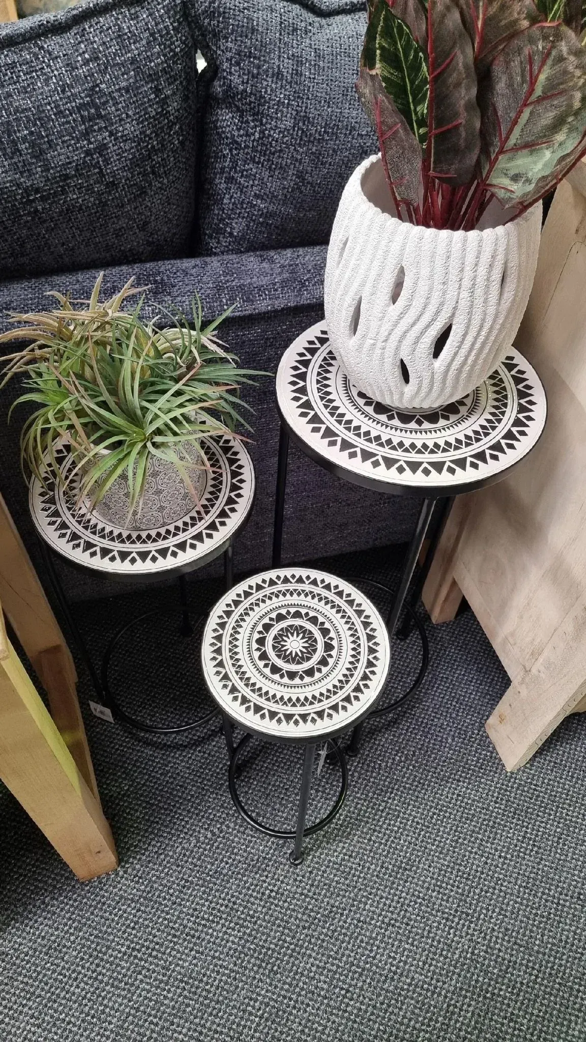 Three small decorative plant stands with potted plants beside a dark gray sofa and beige wall
