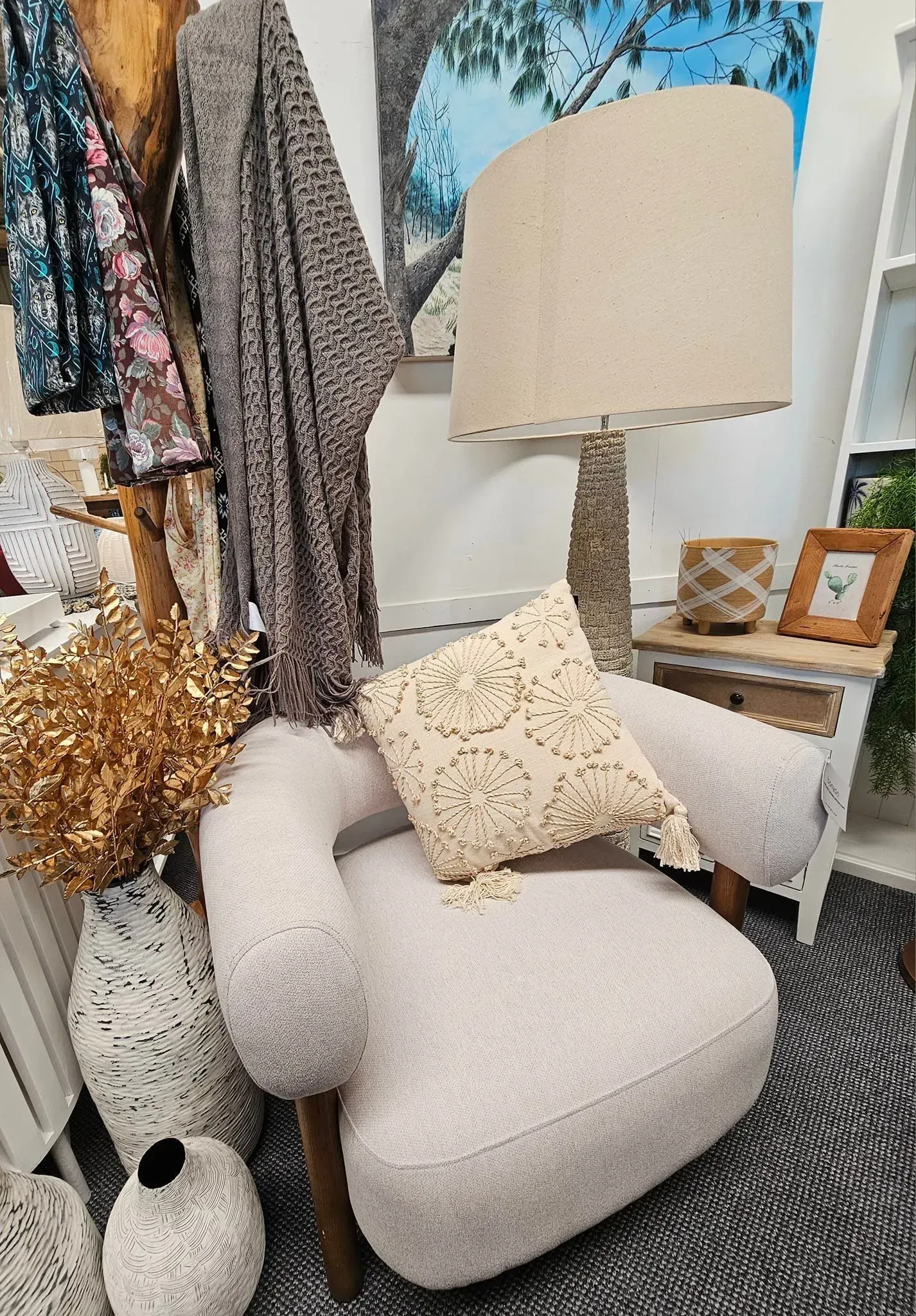 Cozy corner with a cream lounge chair, patterned throw pillow, floor lamp, side table, and hanging clothes underneath