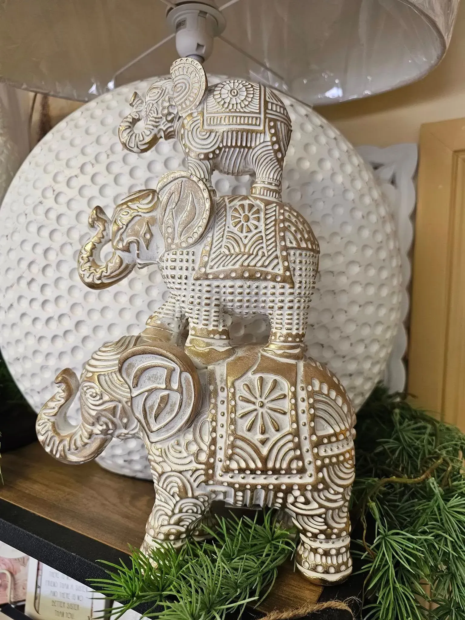 Decorative elephant figurine with gold accents on a wooden shelf, backed by a textured white disc, with greenery below