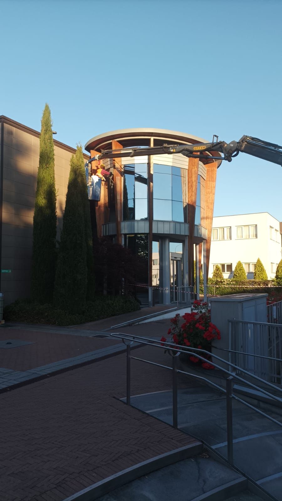 Ingresso di un edificio moderno con pareti in vetro, tetto curvo e fiori rossi accanto a una recinzione metallica al tramonto.
