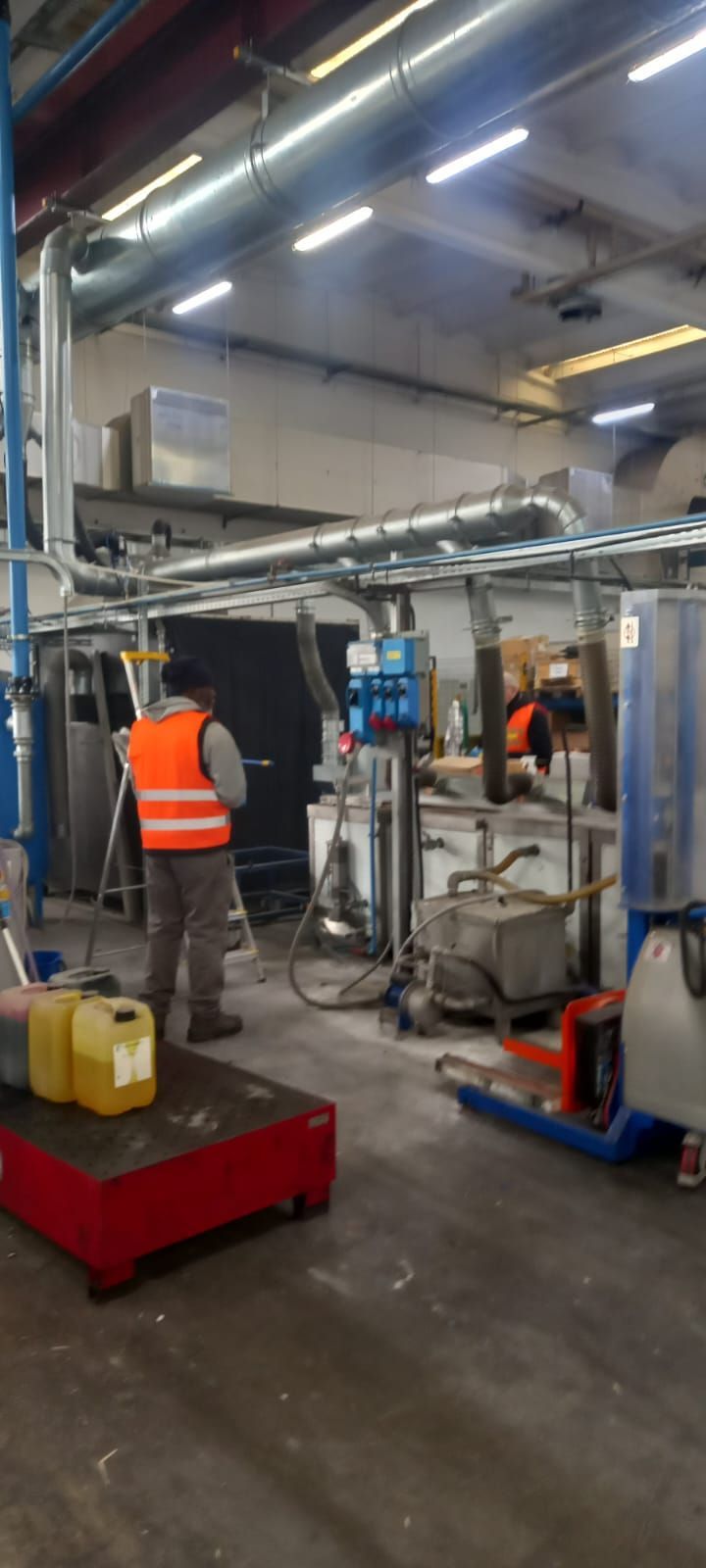 Officina industriale con operai intenti ad assemblare una struttura metallica sotto luminose luci dall'alto.