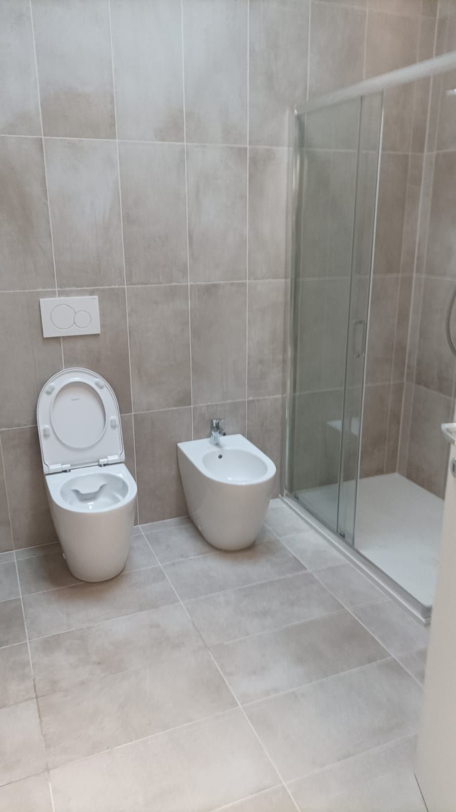 Piccolo bagno piastrellato di beige con WC a vista, bidet e box doccia in vetro.