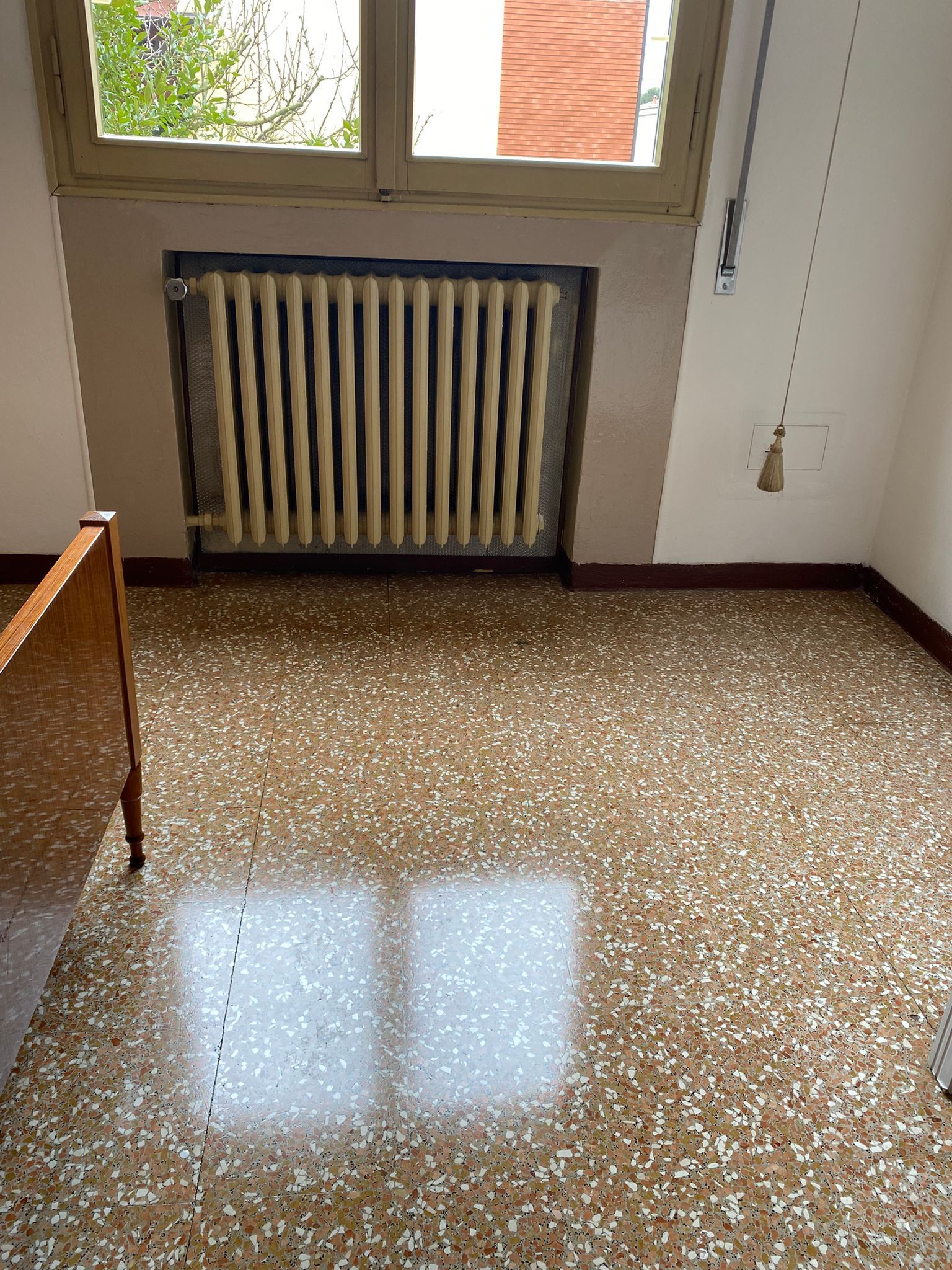 Angolo di una piccola stanza con finestra, termosifone, pavimento in terrazzo e un piano di lavoro in legno.