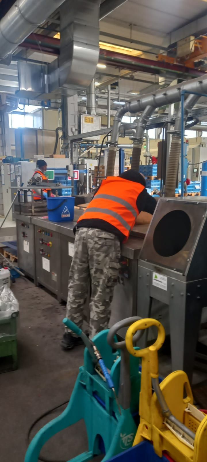 Operaio con giubbotto arancione chino su un macchinario in uno stabilimento industriale con attrezzature e tubi blu.