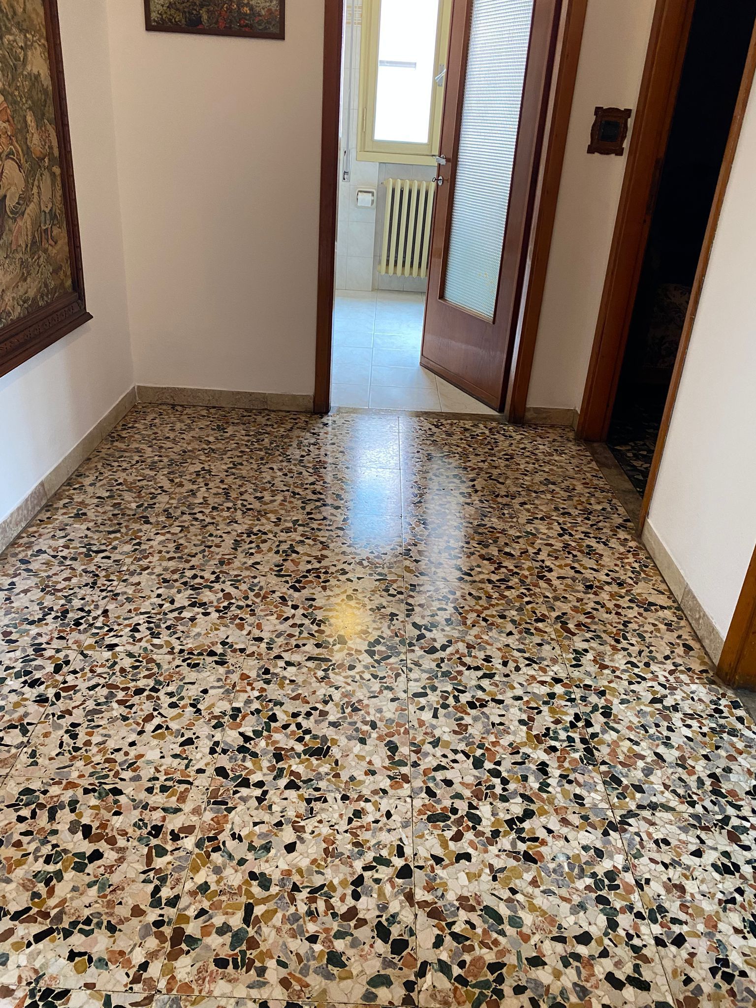 Stretto corridoio con pavimento in terrazzo screziato che conduce a una luminosa porta e al corrimano delle scale.