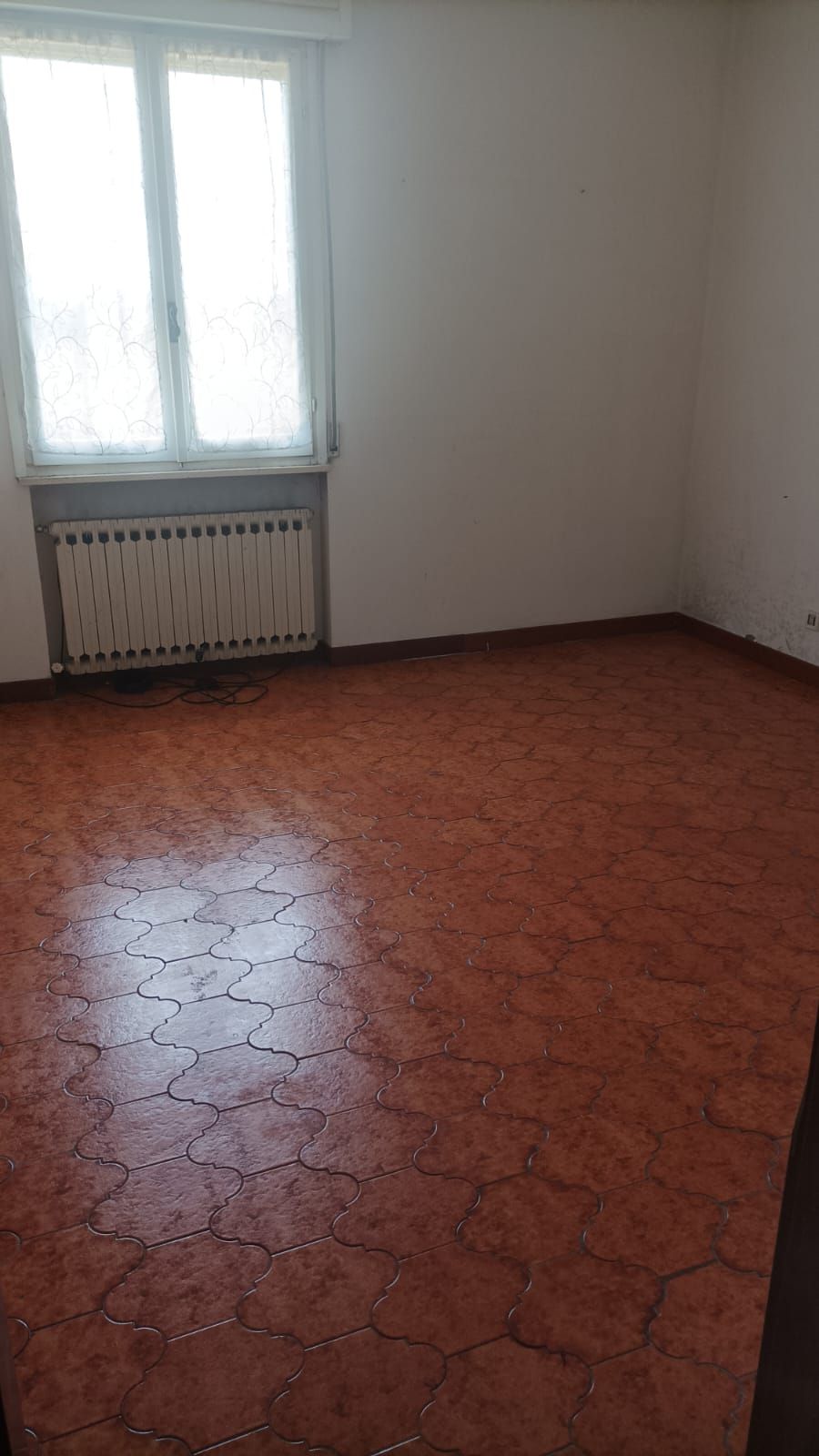 Stanza vuota con pareti bianche, una finestra, un termosifone e un pavimento piastrellato di colore rossastro.