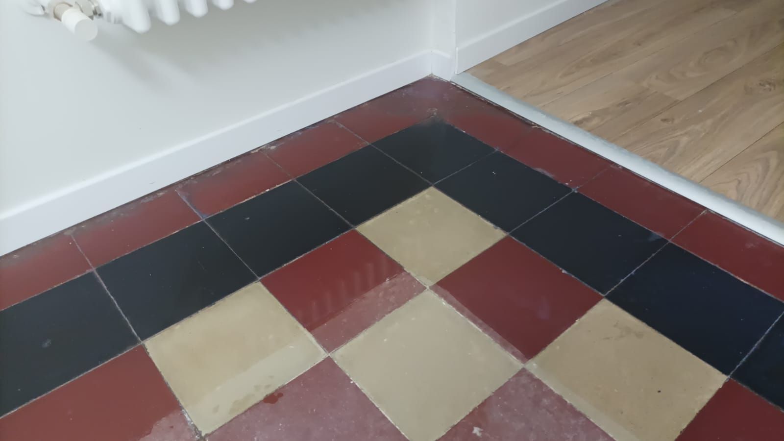 Angolo con pavimento piastrellato di rosso, nero e beige, accanto a una parete bianca e un termosifone.