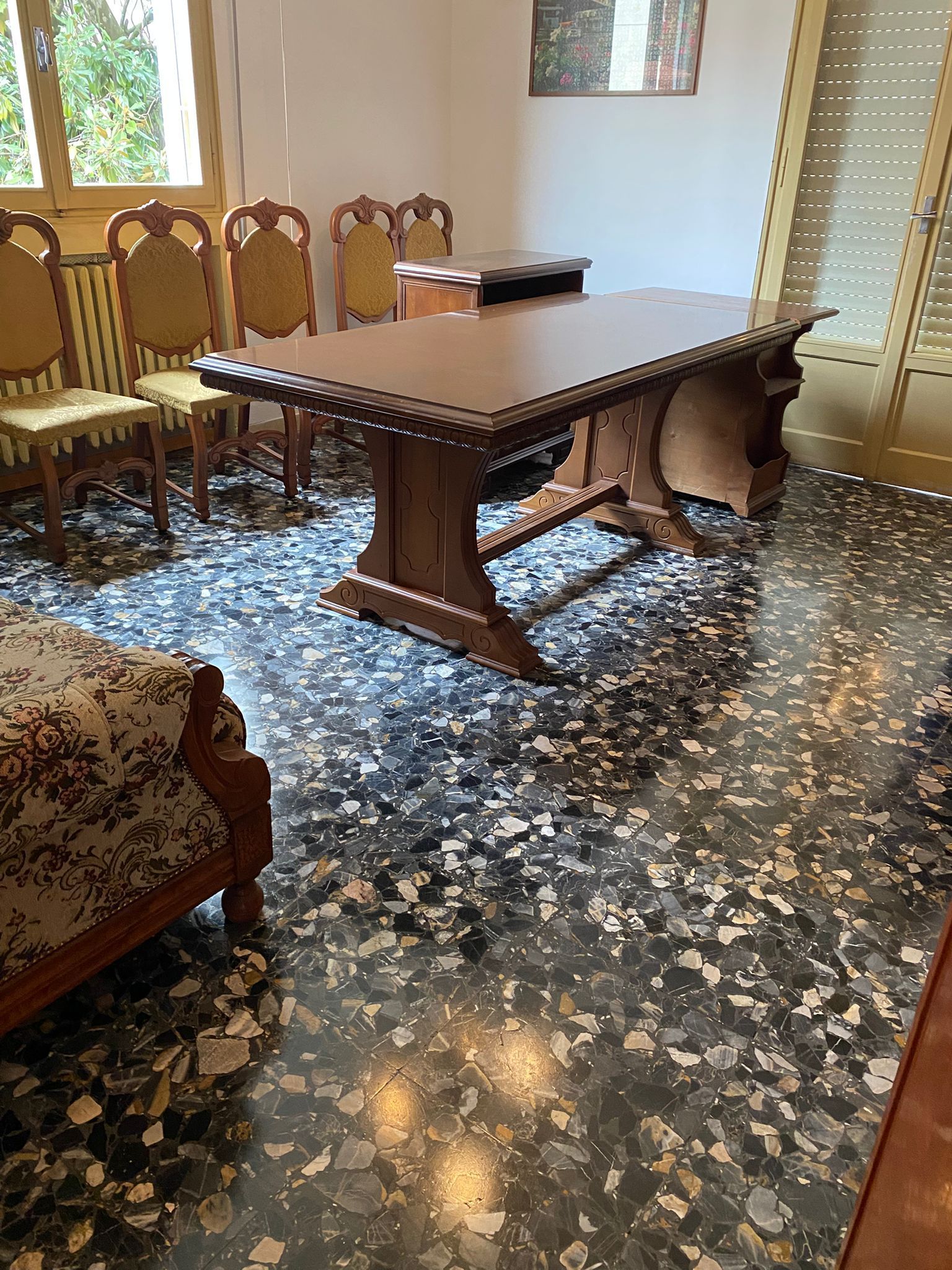 Sala da pranzo con tavolo e sedie in legno su un pavimento piastrellato a motivi geometrici, con finestre luminose e porta a vetri.