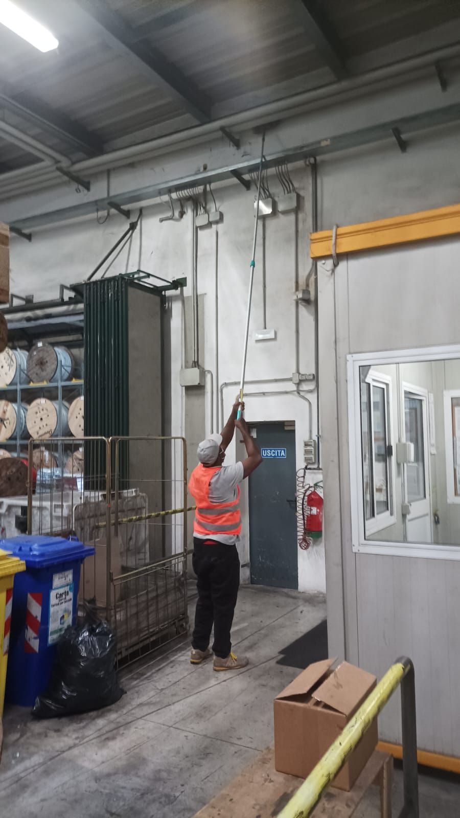 Un operaio con un giubbotto arancione si allunga per ispezionare i tubi in un corridoio di un magazzino industriale.