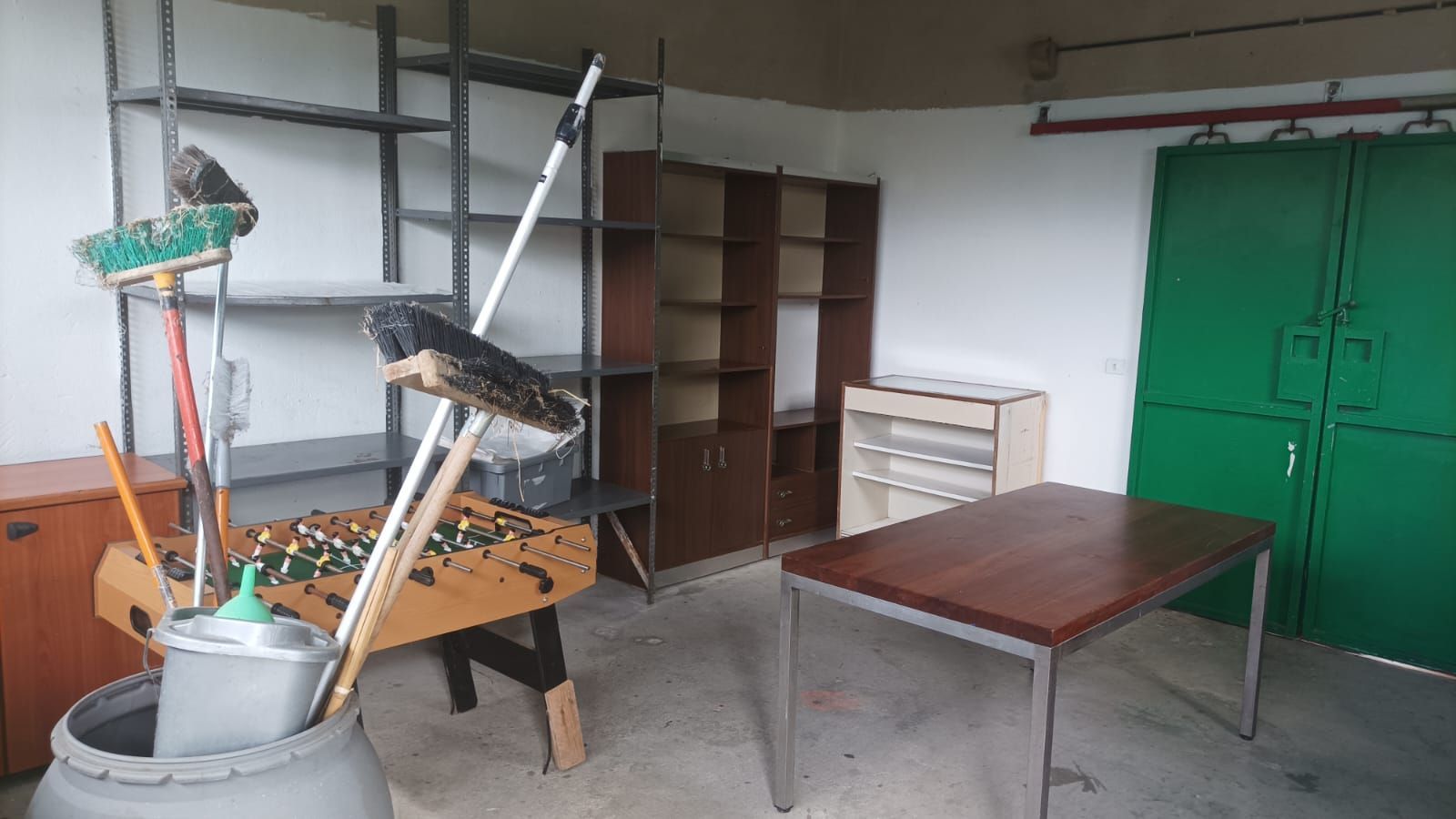 Locale adibito a officina con tavolo metallico, scaffali, armadietto verde e, sulla sinistra, un macchinario su ruote.