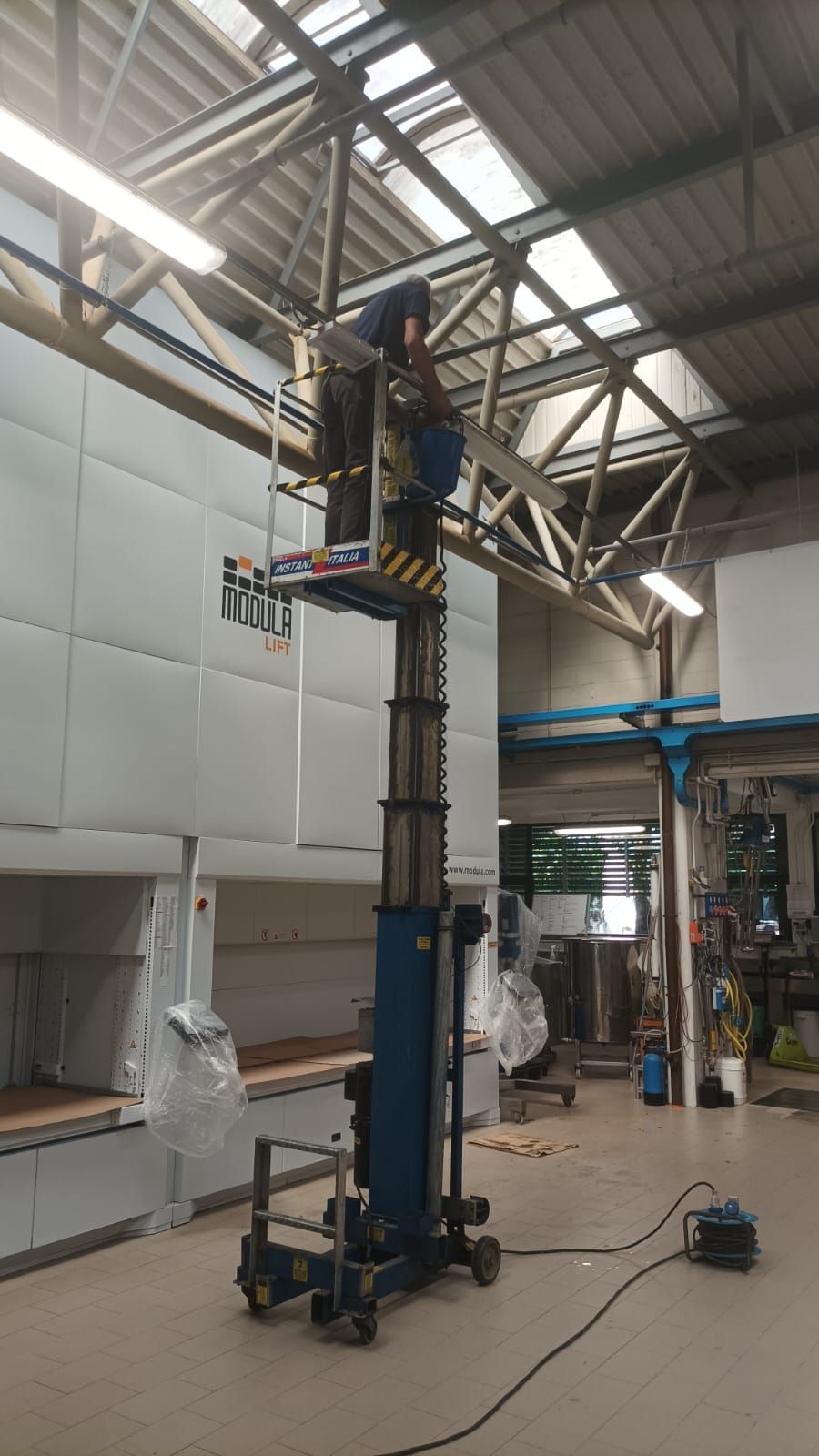 Alto montante blu di un ascensore industriale all'interno di un magazzino, con un operaio su una piattaforma vicino al soffitto.
