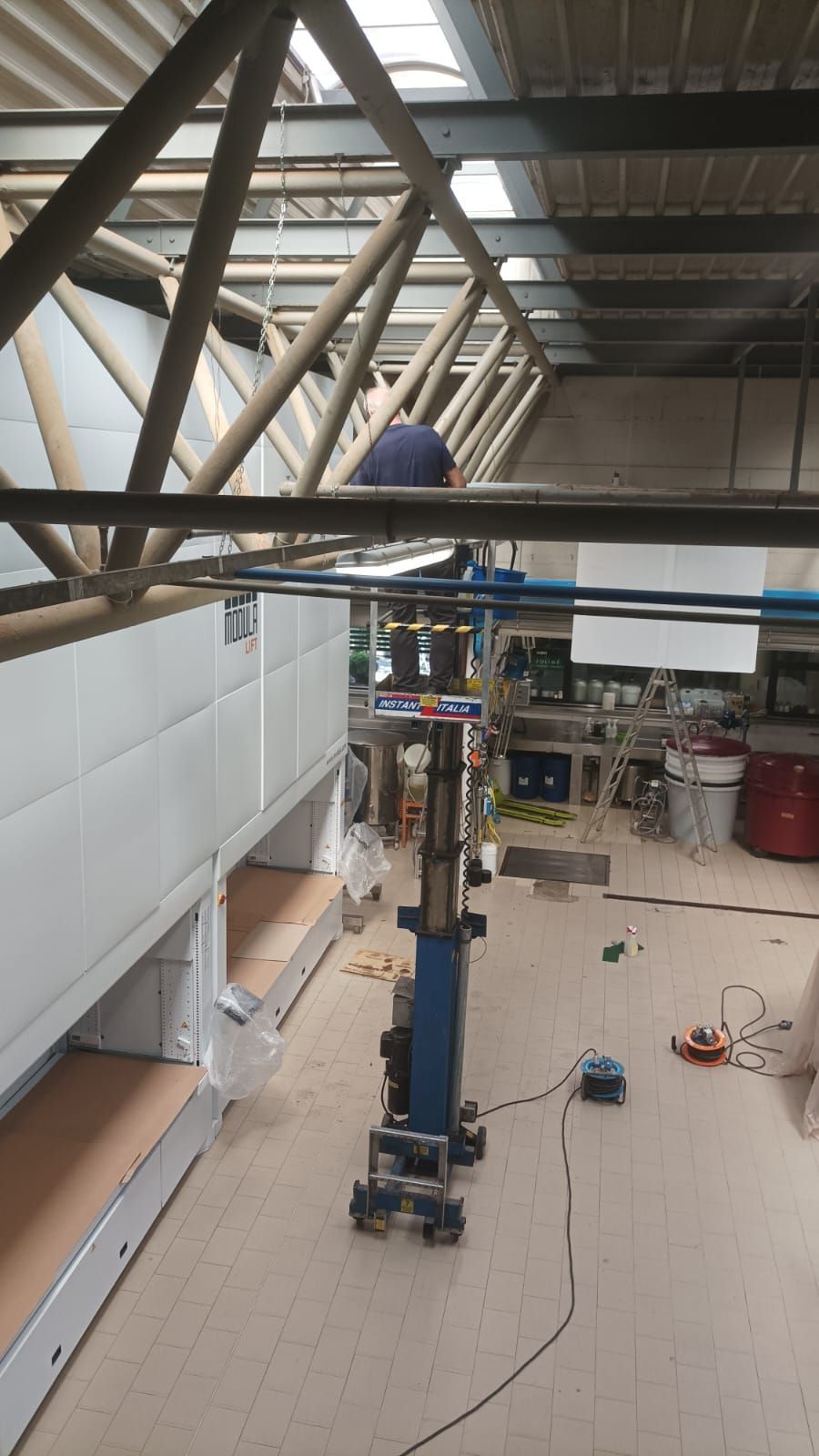 Un operaio su una piattaforma elevatrice blu installa una capriata del soffitto in un magazzino industriale.