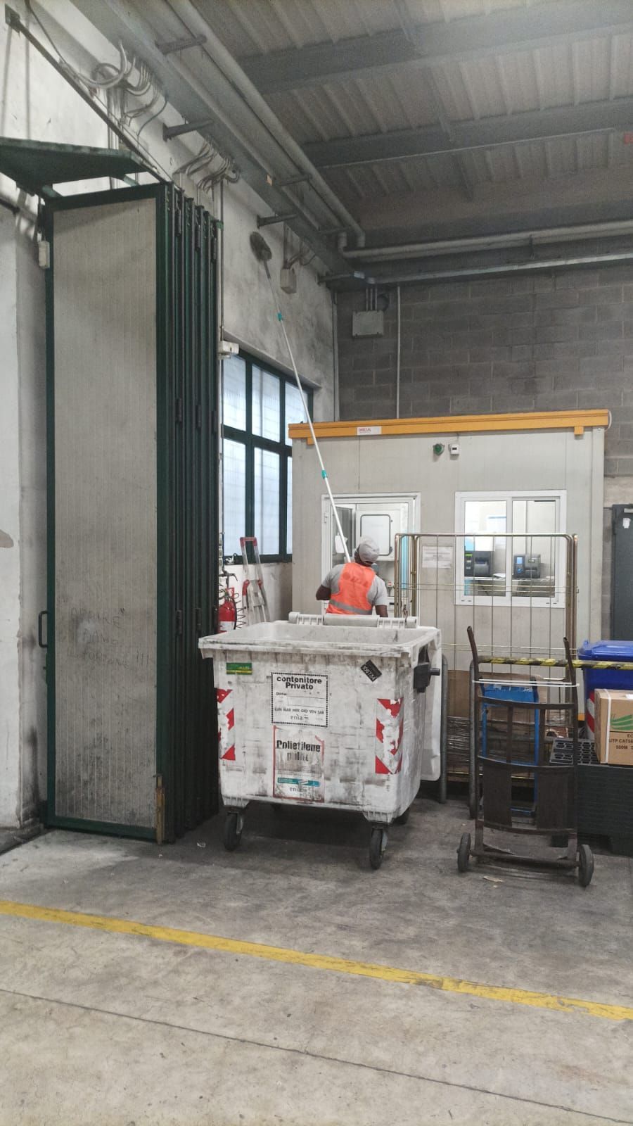 Banchina di carico industriale con carrello per cassonetti, piccolo banco da lavoro e pareti di magazzino in cemento.