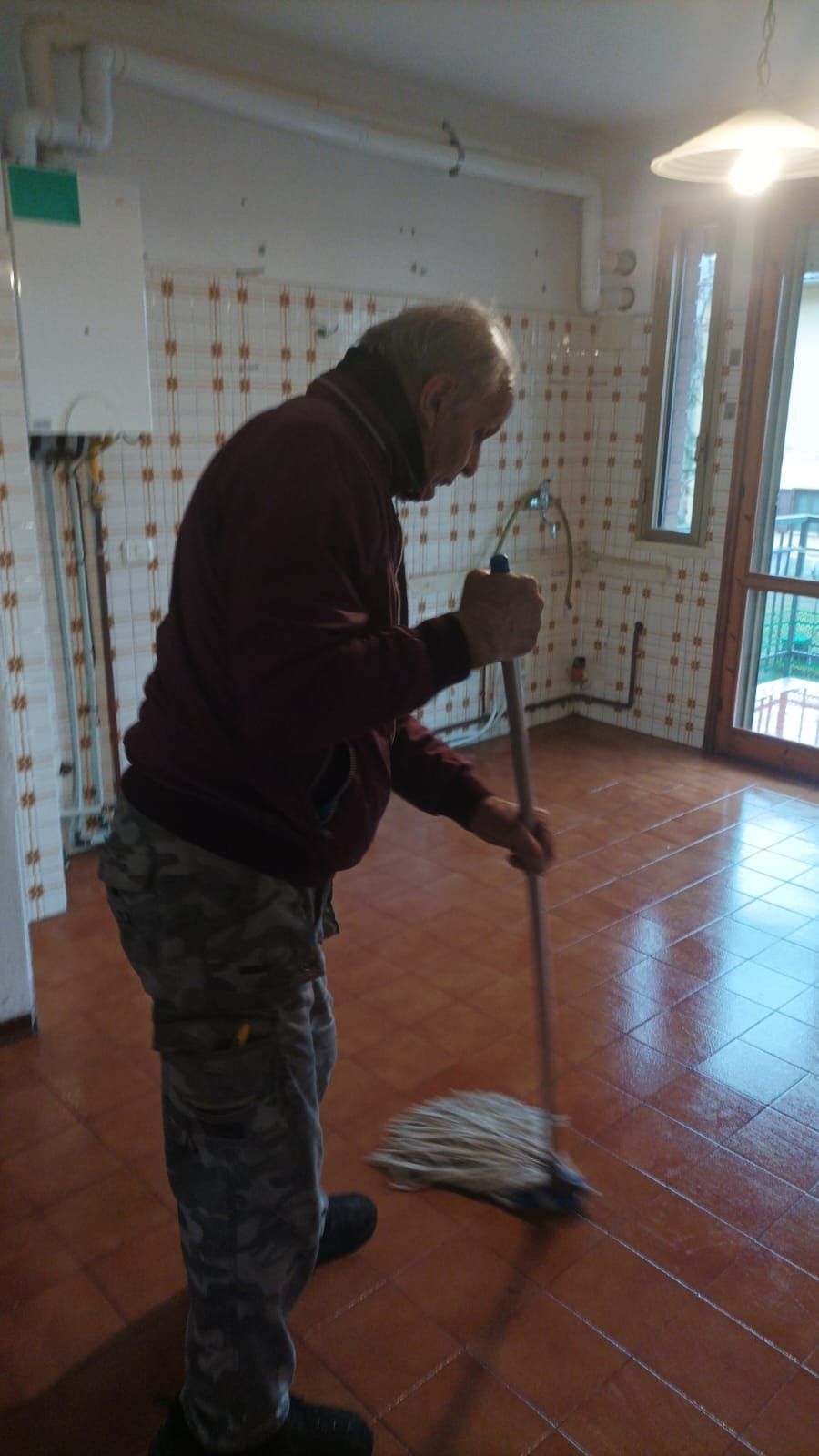 Una persona lava un pavimento piastrellato in una stanza luminosa con porta finestra e balcone.
