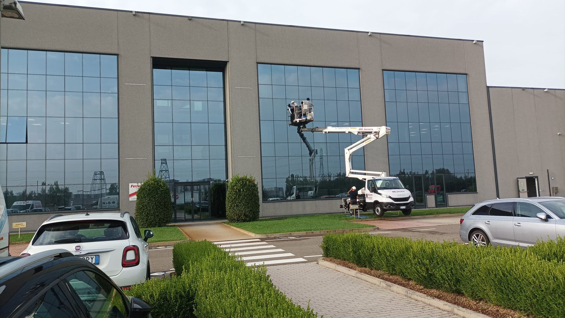 Edificio per uffici con facciata in vetro, con una piattaforma aerea bianca che solleva un operaio, auto parcheggiate e aree verdi antistanti.