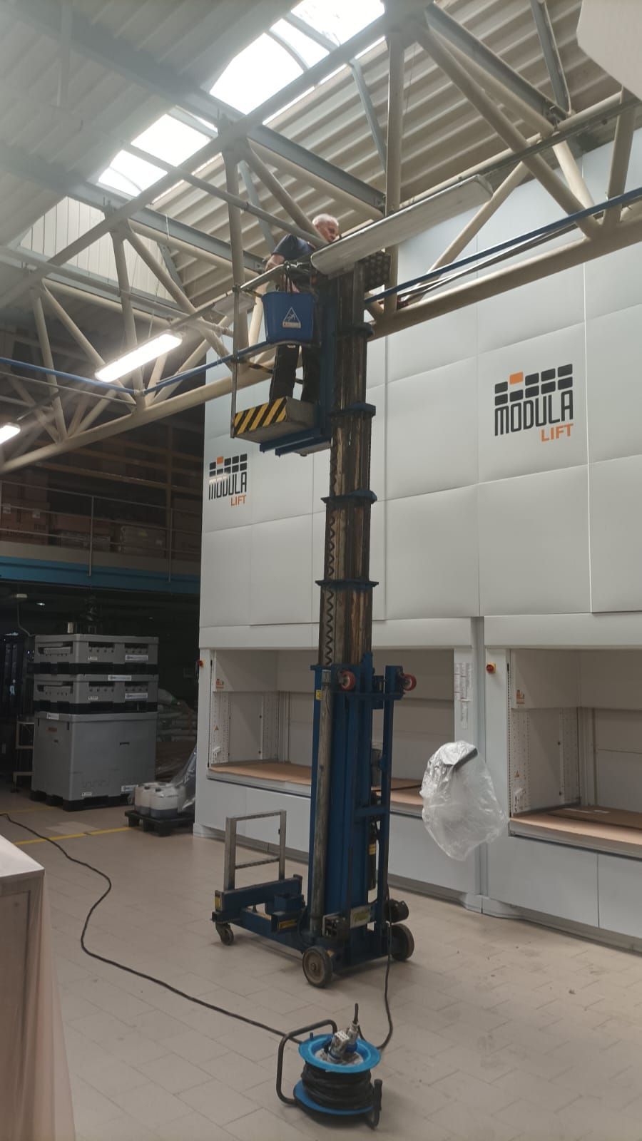In un'officina, un alto elevatore industriale blu si protende verso il soffitto sotto i lucernari.